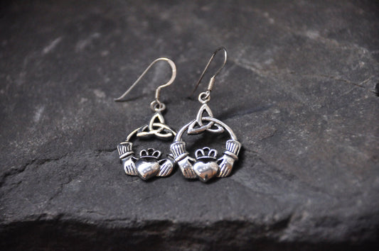 A pair of sterling silver Claddagh earrings with a Triquetra knot design, placed on a dark stone surface.